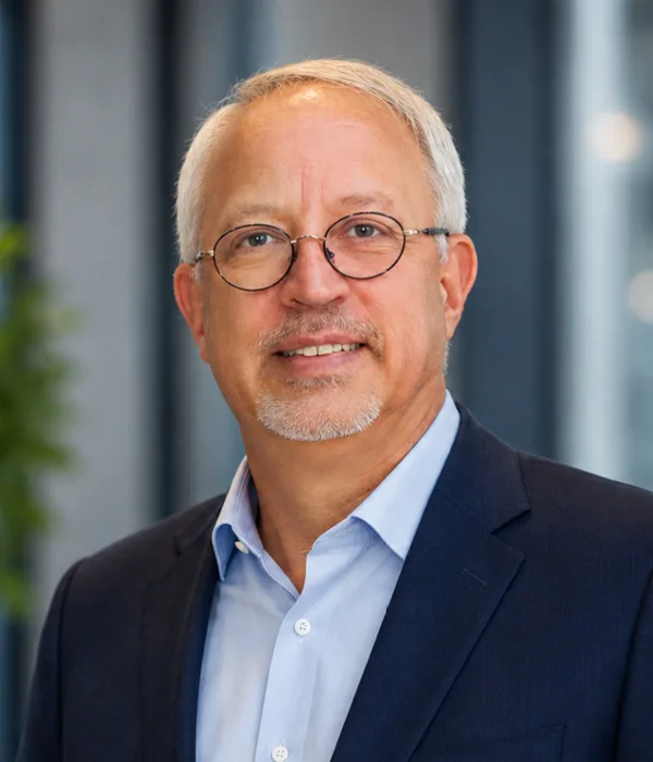 Portrait of an older man with gray hair and round glasses, wearing a navy blazer and light blue shirt, smiling in a modern office setting.