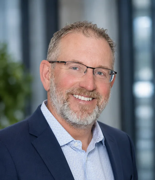 Smiling middle-aged man with glasses and a gray beard, wearing a navy blazer and light blue shirt in an office setting.