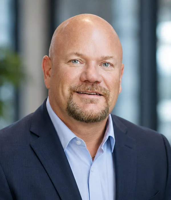 Professional headshot of a bald man in a navy blazer and light blue shirt, smiling in an office setting.
