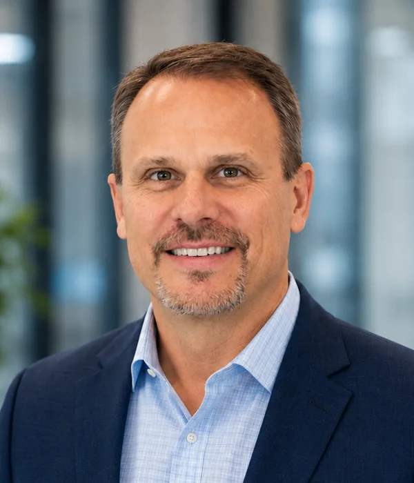 Professional headshot of a smiling man in a navy blazer and light blue shirt against a blurred office background, suitable for business profiles.
