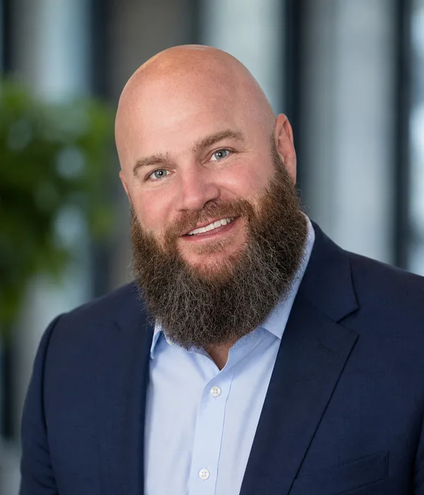 Smiling bald man with a full beard in a navy blazer and light blue shirt, professional headshot.