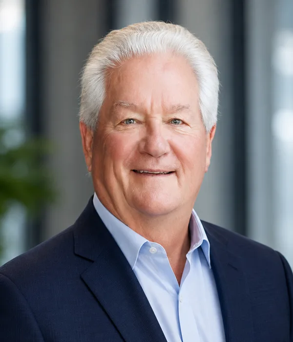 Smiling older man in a navy blazer and light blue shirt, head-and-shoulders portrait in an office setting.