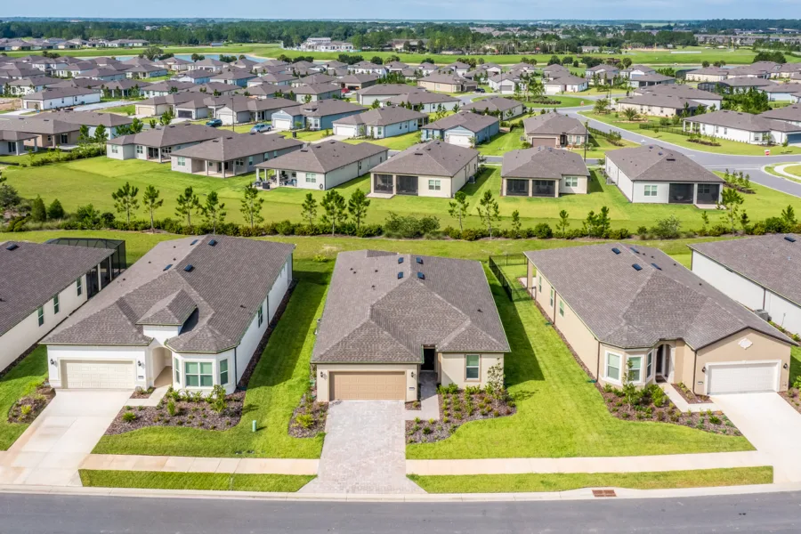 Aerial view of a suburban housing development with rows of single-family homes and green lawns along curved streets.