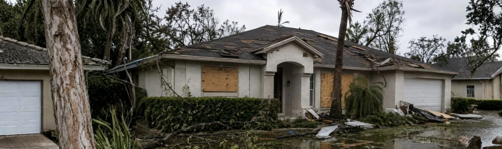 Flooded single-family home with boarded windows and a damaged roof, debris in the yard and water pooling in the driveway area.
