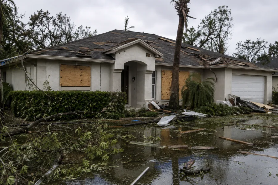 Flooded front yard and a damaged single-story house with boarded windows and scattered debris after a storm.